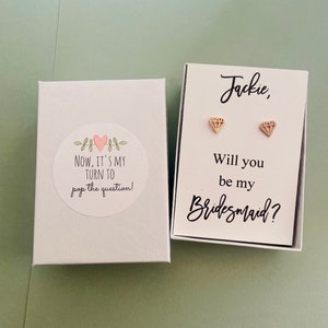 Può includere: Una scatola bianca con un cuore rosa e il testo "NOW, IT'S MY TURN TO pop the question!"  Accanto c'è una scatola bianca con il testo "Jackie, Will you be my Bridesmaid?" e due orecchini a forma di diamante dorati.