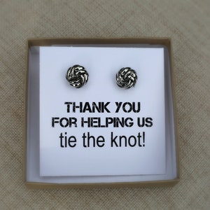 May include: A small gift box containing two silver knot-shaped stud earrings. The earrings are displayed on a white card with the text "THANK YOU FOR HELPING US tie the knot!" printed in black. The box is open, revealing the contents.