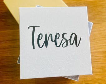 May include: A white box with the name "Teresa" written in black cursive lettering. The box is sitting on a wooden surface.