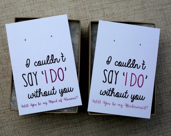 May include: Two white cards with black text and pink accents. The text on the cards reads "I couldn't SAY 'I DO' without you." One card asks "Will you be my Maid of Honour?" and the other asks "Will you be my Bridesmaid?"