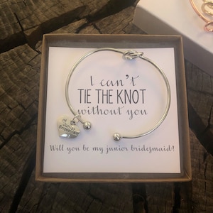 Può includere: Un braccialetto d'argento con un ciondolo a nodo e un ciondolo a forma di cuore che dice "Junior Bridesmaid". Il braccialetto è in una scatola con un biglietto che dice "Non posso fare il nodo senza di te. Vuoi essere la mia damigella d'onore junior?"