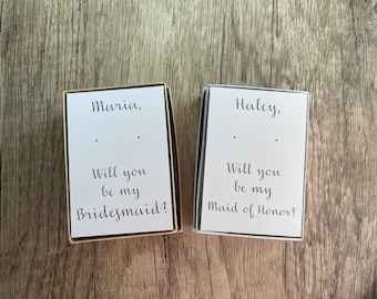 May include: Two small white boxes with black text. The top box says "Maria, Will you be my Bridesmaid?" The bottom box says "Haley, Will you be my Maid of Honor?"