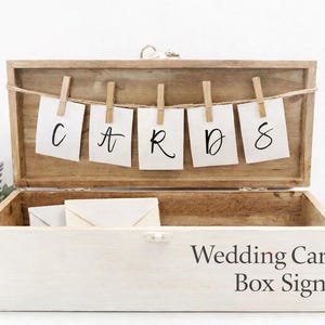Puede incluir: Caja de tarjetas de boda de madera con la palabra "CARDS" mostrada en pequeñas tarjetas suspendidas con pinzas. La caja está abierta, revelando espacio para tarjetas y sobres. La caja está pintada de blanco con el texto "Wedding Card Box Sign".
