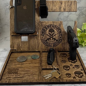 May include: Wooden organizer with compartments for a phone, watch, keys, a knife, and a gun. The organizer has a skull and crossbones design with the text "2nd Amendment" and "We the People".