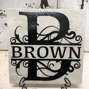 May include: A white ceramic tile with a black letter "B" and the word "BROWN" in black lettering. The letter "B" has a decorative flourish design. The tile is on a black metal stand.