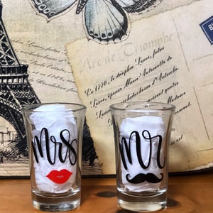 May include: Two clear glass shot glasses with black lettering. One glass says "Mrs." with a red lipstick mark. The other glass says "Mr." with a black mustache.