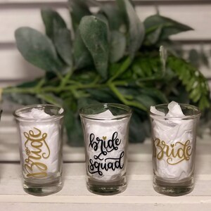 May include: Five clear glass shot glasses with black and gold lettering. The glasses say "Bride Team", "Bride", "Bride Squad", "Bride", and "Bride Tribe".