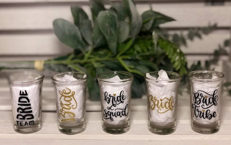 May include: Five clear glass shot glasses with black and gold lettering. The glasses say "Bride Team", "Bride", "Bride Squad", "Bride", and "Bride Tribe".