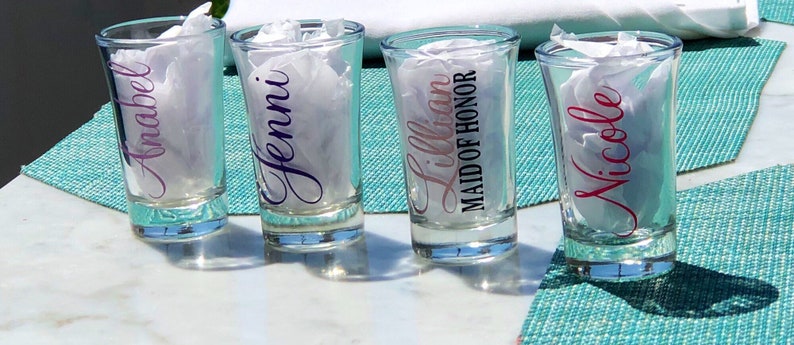 May include: Four clear glass shot glasses with personalized names and titles, including "Anabel", "Jenni", "Lillian Maid of Honor", and "Nicole".