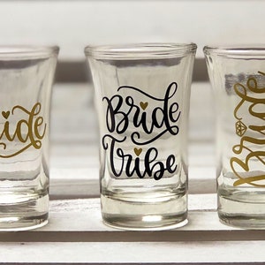 May include: Five clear glass shot glasses with black and gold lettering. The glasses say "bride squad", "bride", "bride tribe", "bride", and "team bride".