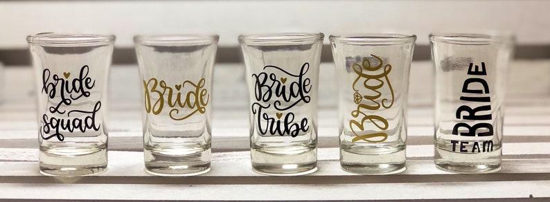 May include: Five clear glass shot glasses with black and gold lettering. The glasses say "bride squad", "bride", "bride tribe", "bride", and "team bride".