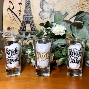 May include: Three clear glass shot glasses with black lettering and gold hearts. The glasses say "Bride Squad", "Bride", and "Bride Tribe".