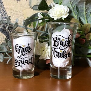 May include: Two clear glass shot glasses with black calligraphy that reads "bride squad" and "bride tribe".  The text is accented with gold hearts.