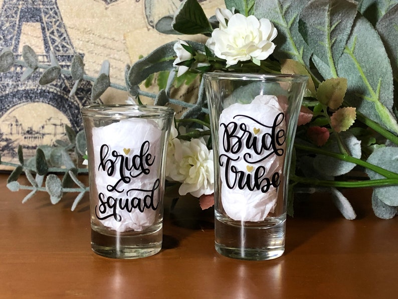 May include: Two clear glass shot glasses with black calligraphy that reads "bride squad" and "bride tribe".  The text is accented with gold hearts.