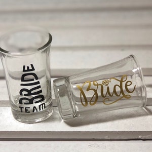 May include: Two clear glass shot glasses with black and gold lettering. One glass says "Team Bride" and the other says "Bride" with a diamond graphic.