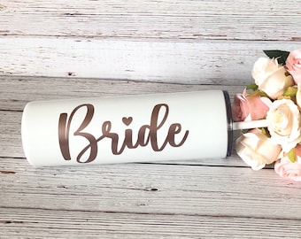 May include: White stainless steel tumbler with a rose gold 'Bride' inscription and a small heart. The tumbler is on a wooden surface with a bouquet of white and pink roses in the background.