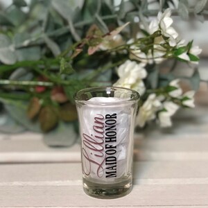 May include: A clear glass shot glass with a black and white design that says "Lillian Maid of Honor" in a cursive font. The text is in a pink color.