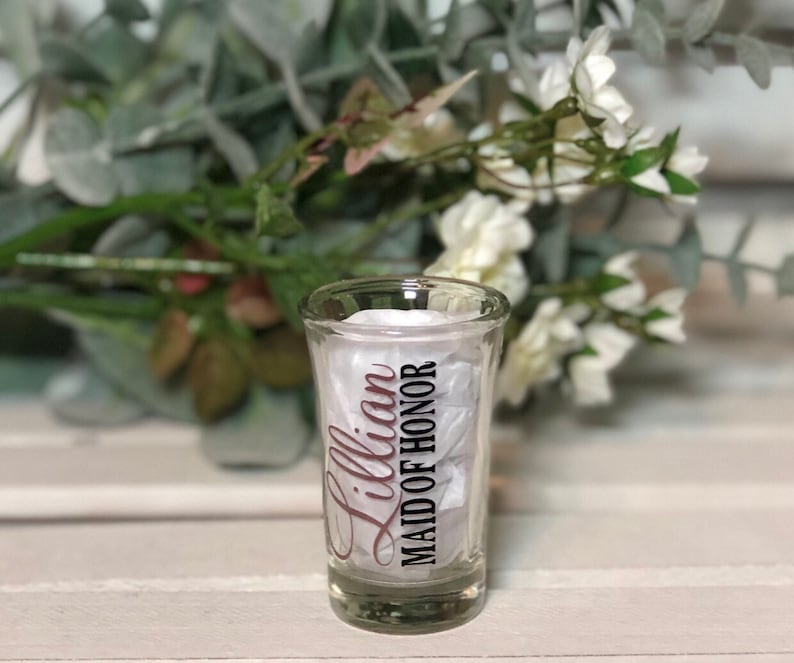 May include: A clear glass shot glass with a black and white design that says "Lillian Maid of Honor" in a cursive font. The text is in a pink color.