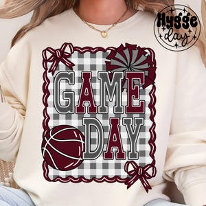 Op de afbeelding: Crèmekleurige sweatshirt met een 'GAME DAY' opdruk. Het ontwerp toont een basketbal, een cheerleader pompom en strikken, allemaal in bordeauxrood en grijs tegen een geruite achtergrond. De tekst is grijs omlijnd en bordeauxrood gevuld.