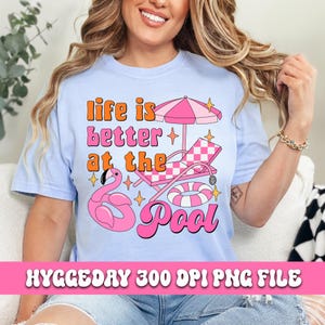Pode incluir: Uma camiseta azul clara com o texto "Life is better at the pool" em letras rosa e laranja. O design tamb&eacute;m inclui um flamingo rosa, uma cadeira de praia listrada de rosa e branco, um guarda-sol listrado de rosa e branco e um anel infl&aacute;vel rosa.