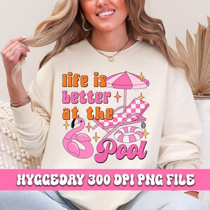 Pode incluir: Uma camisola de cor creme com o texto "life is better at the pool" em rosa, laranja e branco. O design inclui um flutuador de flamingo rosa, uma cadeira de praia listrada rosa e branca e um guarda-sol listrado rosa e branco.