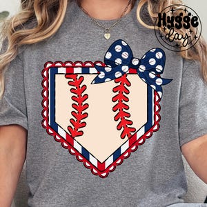 Può includere: T-shirt grigia con un design a tema baseball. Il design include una forma di base di baseball con accenti rossi e blu, un centro color crema e un fiocco blu navy a pois bianchi. Il testo "Hygge day" si trova nell'angolo in alto a destra.