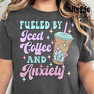 May include: A dark gray t-shirt with the text "Fueled by Iced Coffee and Anxiety" in a retro font. The text is in shades of blue, purple, and pink. A cartoon drawing of a glass of iced coffee with ice cubes is to the right of the text.