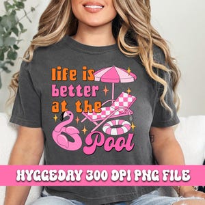 Pode incluir: Uma camiseta cinza com o texto "Life is better at the pool" em letras rosa e laranja. O design tamb&eacute;m inclui um flamingo rosa, uma espregui&ccedil;adeira xadrez rosa e branca, um flutuador de piscina rosa e branco e estrelas.