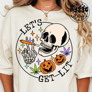 Può includere: T-shirt color crema con grafica circolare raffigurante un teschio, una mano scheletrica che tiene una sigaretta accesa e due zucche di Halloween. Il testo "LET'S GET LIT" è stampato attorno al disegno, con stelle e foglie di cannabis.