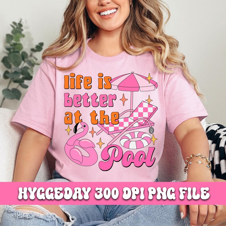Pode incluir: Camiseta rosa com o texto "life is better at the pool" em uma fonte retr&ocirc;. O design inclui um flamingo rosa, um guarda-sol de praia listrado rosa e branco, uma espregui&ccedil;adeira xadrez rosa e branca e um flutuador de piscina listrado rosa e branco.