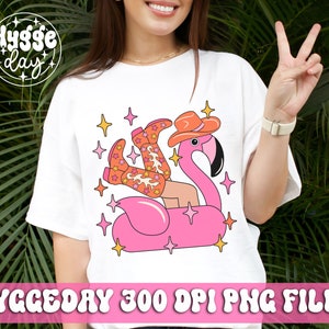 May include: A white t-shirt with a pink flamingo and a person wearing cowboy boots and a cowboy hat on a pink inflatable float. The design includes stars and the text "Hygge Day".