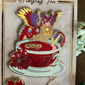Peut inclure: Une &eacute;pingle en &eacute;mail repr&eacute;sentant un dragon rouge, jaune et violet avec une cha&icirc;ne dor&eacute;e attach&eacute;e &agrave; une fleur d'hibiscus rouge. Le dragon tient une tranche d'orange et est assis dans une tasse &agrave; th&eacute; remplie de liquide rouge. L'&eacute;pingle indique "Hibiscus Tea" sur le fond.