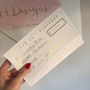 May include: A white rectangular gift cheque with gold foil lettering. The text reads HAPPY 21ST BIRTHDAY, TO Our darling Charlie, AMOUNT Five thousand, £ 5,000, DATE 16/07/05, and LOVE FROM Mum & Dad. The cheque features a double-line border.