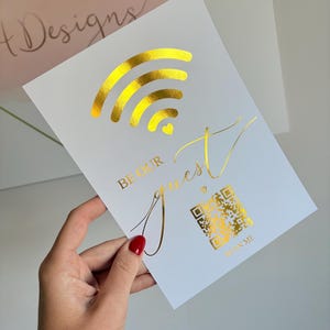 May include: A white rectangular sign featuring gold foil accents. The design includes a gold Wi-Fi symbol with a small heart, the text "BE OUR guest" in a mix of block and script fonts, a gold QR code, and the text "SCAN ME" below the code.