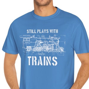 Può includere: Una maglietta blu chiaro con un disegno bianco di un treno e il testo "Still Plays With Trains".