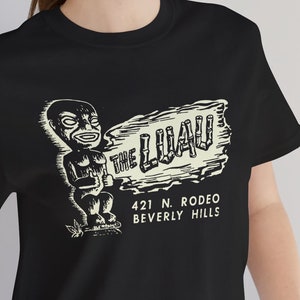 Könnte beinhalten: Schwarzes T-Shirt mit einem weißen Aufdruck eines Tiki-Figuren, der ein Schild mit der Aufschrift "The Luau" hält, darunter die Adresse "421 N. Rodeo Beverly Hills".