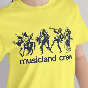 Op de afbeelding: Een felgeel T-shirt met een marineblauwe afbeelding van muzikanten en de tekst "musicland crew". Het ontwerp toont figuren die instrumenten bespelen. Het T-shirt is gemaakt van een zacht materiaal.