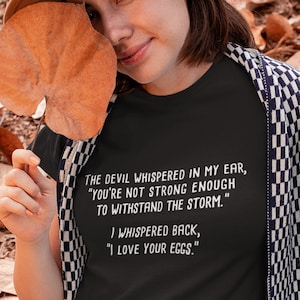 May include: A black t-shirt with white text that reads "The devil whispered in my ear, "You're not strong enough to withstand the storm." I whispered back, "I love your eggs."