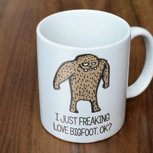 May include: White ceramic mug featuring a cartoon Bigfoot illustration in brown and black. The mug has the text "I JUST FREAKING LOVE BIGFOOT, OK?" in black. The mug has a curved handle and is suitable for hot or cold beverages.