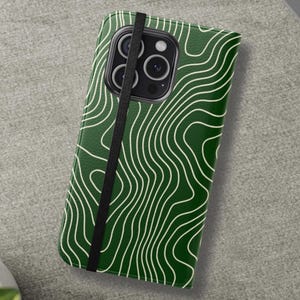 May include: A green phone case with a white topographic pattern. The case is closed with a black elastic band. The phone case is on a gray surface, with a laptop, pen, and earbuds in the background.