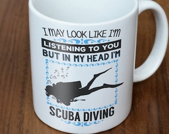 Puede incluir: Taza de cerámica blanca con una silueta de buzo negra y el texto "I MAY LOOK LIKE I'M LISTENING TO YOU BUT IN MY HEAD I'M SCUBA DIVING" en negro y azul. La taza tiene un asa curvada.