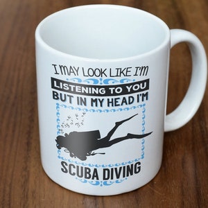May include: White ceramic mug with a black scuba diver silhouette and the text "I MAY LOOK LIKE I'M LISTENING TO YOU BUT IN MY HEAD I'M SCUBA DIVING" in black and blue. The mug has a curved handle.