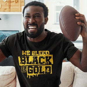 以下が含まれることがあります： 「WE BLEED BLACK & GOLD」の文字が黄色でプリントされた黒いTシャツ。Tシャツを着た人が茶色のフットボールを持ち、祝っています。その人はベージュのソファに座っています。