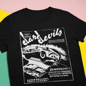 Puede incluir: Camiseta negra con un diseño gráfico retro. El diseño presenta la palabra "Daredevils" e imágenes de coches clásicos en acción. El gráfico también incluye el texto "28 Hair Raising & Death Defying Stunt Events" y "Under Brilliant Track Lighting!"