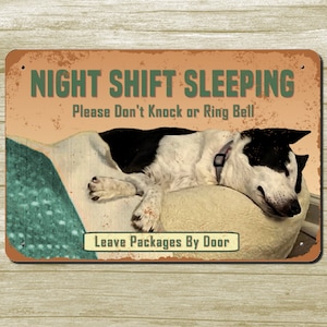 May include: A metal sign with a rustic orange background and green text that reads "Night Shift Sleeping. Please Don't Knock or Ring Bell. Leave Packages By Door". A black and white dog is sleeping on a white pillow with a green blanket.
