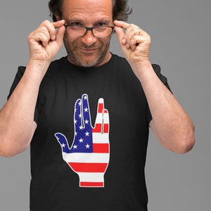 Peut inclure: T-shirt noir avec un motif graphique d'une main en signe de salut vulcain, intégrant le design du drapeau américain. Le motif de la main présente des étoiles bleues et des rayures rouges et blanches. Le t-shirt est porté par une personne.
