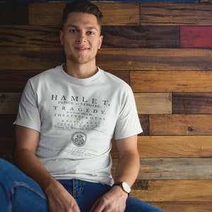Op de afbeelding: Een wit T-shirt met een zwarte print van het toneelstuk Hamlet, Prins van Denemarken: Een tragedie. De print bevat de tekst "Theatres Royal Drury Lane Covent Garden" en "Geschreven door William Shakespeare".