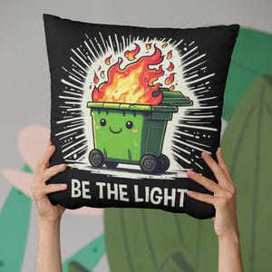 May include: Black decorative pillow featuring a cartoon green trash bin on fire with a smiling face. The pillow has the text "BE THE LIGHT" in white. The background has white radiating lines.