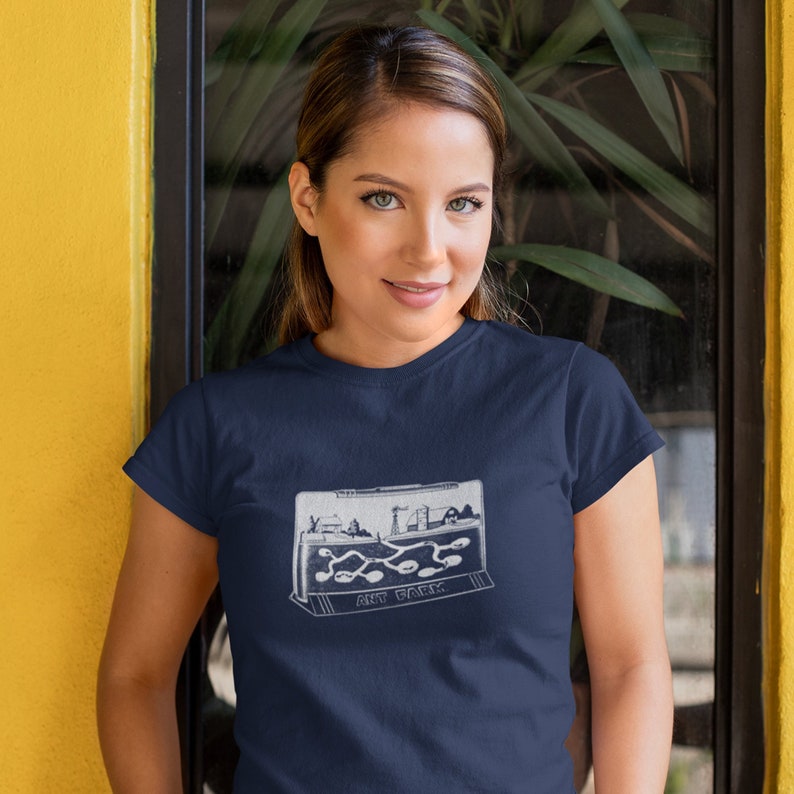 May include: A navy blue t-shirt with a white graphic of an ant farm. The graphic shows a clear plastic ant farm with ants crawling through tunnels. The text "ANT FARM" is printed below the graphic.