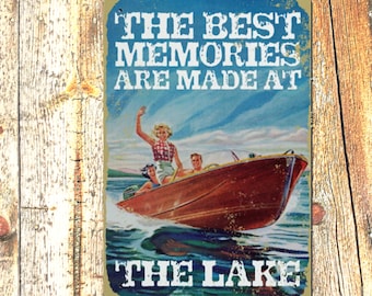 May include: A vintage-style metal sign with a blue background and white text that reads "The Best Memories Are Made At The Lake". The sign features a retro illustration of a boat with three people on it, cruising on a lake.